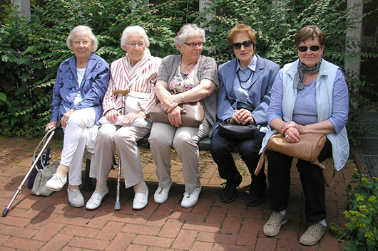 Fnf Teilnehmerinnen des Seniorenausflugs sitzen vor Fahrtbeginn auf der Bank vor der Trinitatiskirche.