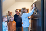 Eltern und Geschwister sehen einem Mdchen zu, das auf einer Leiter steht und den Segensaufkleber der diesjhrigen Sternsinger-Aktion befestigt.