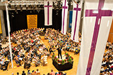 Blick auf die Bühne der Stadthalle in Stadtallendorf, auf der Bläserinnen und Bläser aus zahlreichen Posaunenchören im Halbkreis sitzend ihre Instrumente spielen.