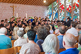 Blick durch die Zuschauerreihen in Richtung Altarraum, wo mehr als vierzig Musikerinnen und Musiker in Aktion zu sehen sind.